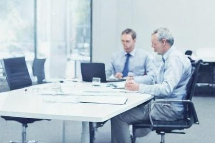 Zwei Männer in Anzügen sitzen an einem Tisch mit Laptops und Notizblättern in einem Besprechungsraum.