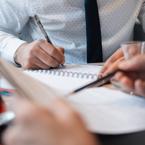 Hände, die in einem Notizbuch schreiben, mit Stiften und Getränken auf dem Tisch.